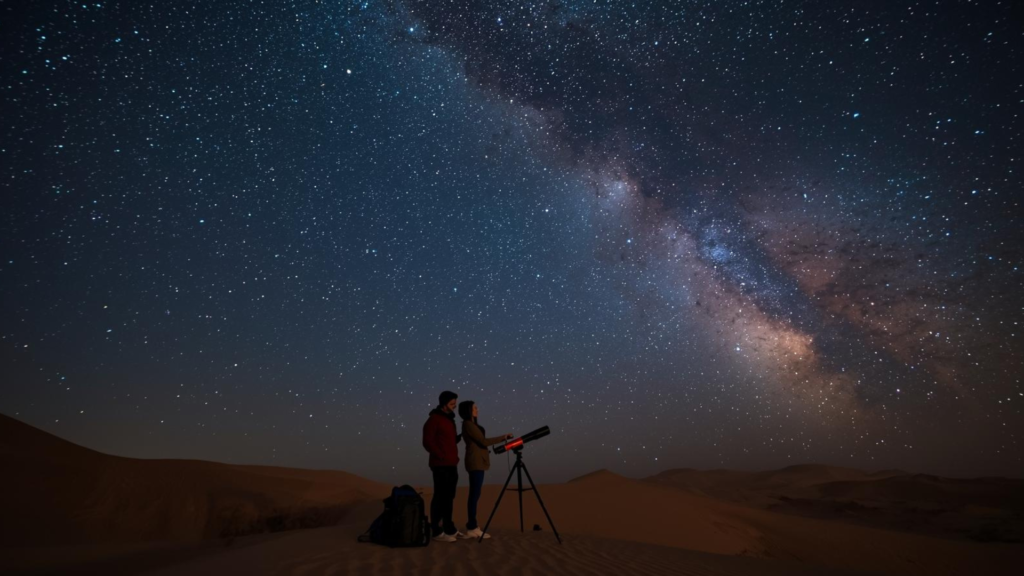 Duas pessoas sentadas no deserto, contemplando um céu estrelado e vasto.